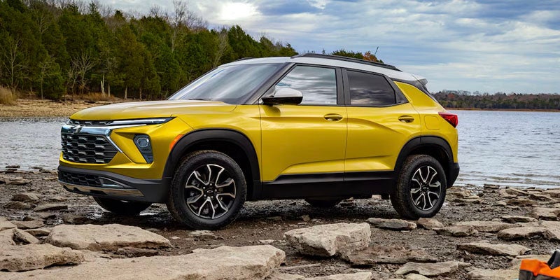 Yellow compact SUV parked on rocky terrain by a lake, with trees and cloudy skies in the background.