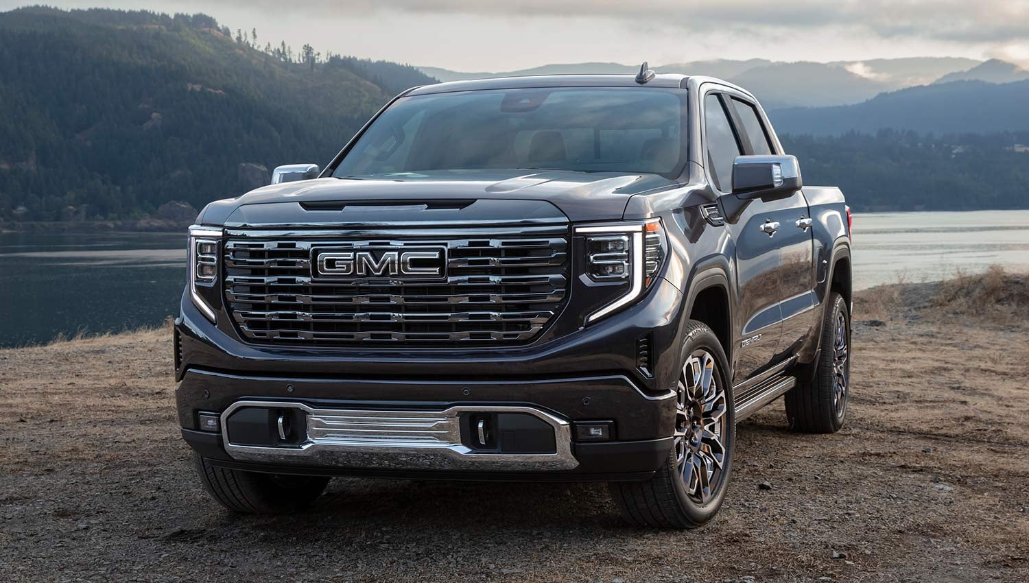 Black GMC pickup truck parked on a dirt road near a lake with mountains in the background.