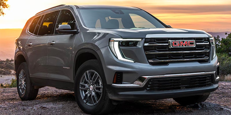A gray 2024 GMC Acadia SUV parked on a rocky terrain at sunset, showcasing its bold front grille and LED headlights.