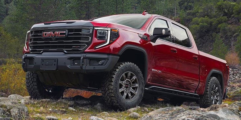 Red GMC Sierra AT4X pickup truck parked on a rocky terrain in a forested area, showcasing its rugged off-road design and bold front grille.