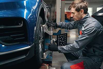 Auto mechanic performing wheel alignment on a blue vehicle using specialized equipment in a service garage.
