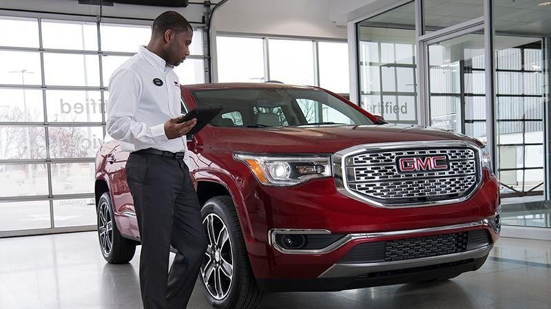 Salesperson holding a tablet while inspecting a red GMC SUV inside a brightly lit car dealership showroom.