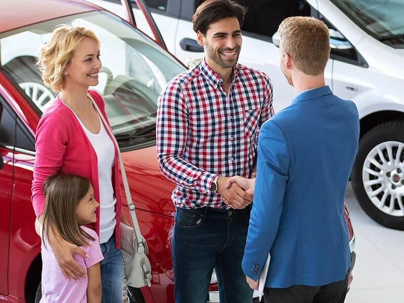 Smiling man shaking hands with a car salesman inside a dealership, with a woman and a young girl standing beside him near a red car.
