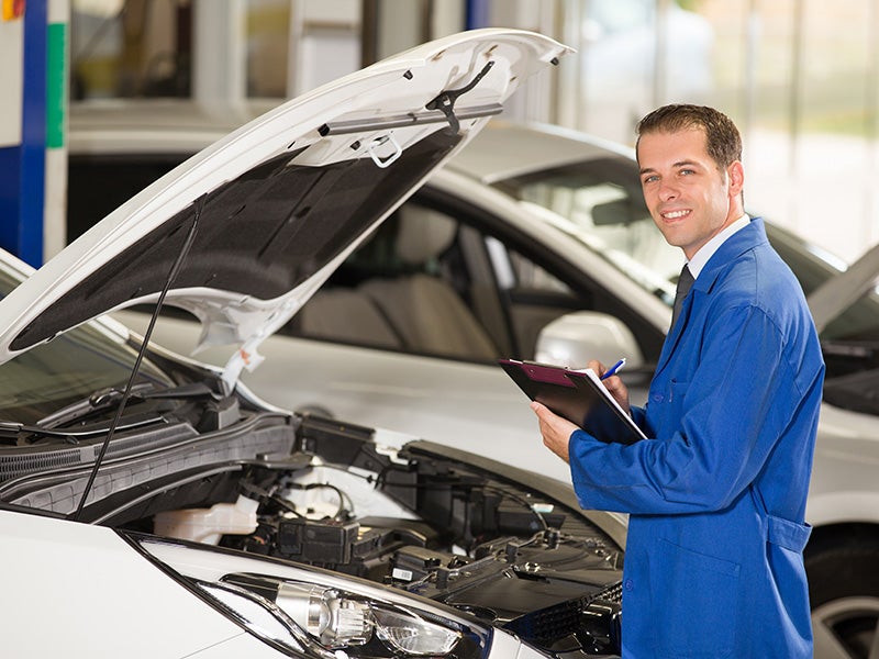 Smiling mechanic in a blue uniform holding a clipboard while inspecting a car with its hood open in an auto repair shop.